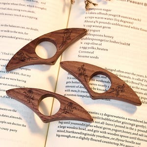 May include: Three wooden book page holders with a floral design are displayed on a book page. The holders are made of dark brown wood and have a circular opening. The book page has text and a recipe for French Peasant Bread.