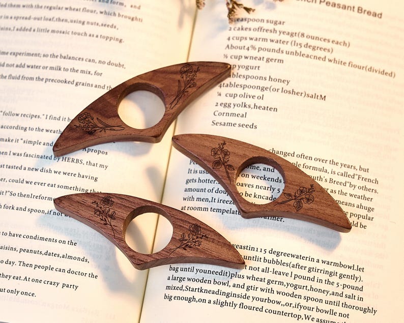May include: Three wooden book page holders with a floral design are displayed on a book page. The holders are made of dark brown wood and have a circular opening. The book page has text and a recipe for French Peasant Bread.