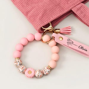 May include: A pink beaded bracelet with floral details and a matching keychain. The keychain has a tassel and the name "Olivia" printed on it. The bracelet features large pink beads and decorative beads with a floral pattern. A pink tote bag is in the background.