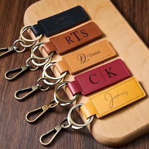 Può includere: Una collezione di portachiavi in pelle personalizzati in vari colori, tra cui nero, marrone, beige, bordeaux e giallo. Ogni portachiavi presenta un anello e un moschettone in metallo, con iniziali o nomi incisi. I portachiavi sono disposti su un tagliere di legno.