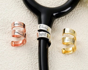 May include: Three personalised rings in copper, silver, and gold. The copper ring is engraved with "Amelia" and an infinity heart symbol. The silver ring has "Olivia" and a floral design. The gold ring is inscribed with "William" and a heartbeat symbol.
