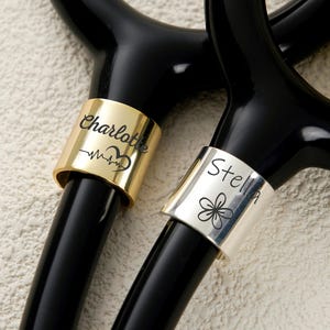 Può includere: Primo piano di due stetoscopi neri con anelli personalizzati. Un anello è dorato con "Charlotte" e un disegno a battito cardiaco. L'altro è argentato con "Stella" e un fiore.