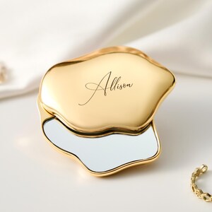 May include: A gold-colored compact mirror with an organic, irregular shape. The mirror is open, revealing a reflective surface. The name "Allison" is elegantly scripted on the top. A gold ring with a geometric design is also present.