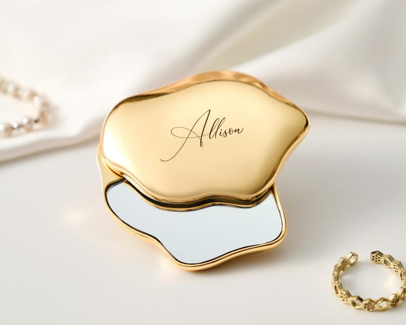 May include: A gold-colored compact mirror with an organic, irregular shape. The mirror is open, revealing a reflective surface. The name "Allison" is elegantly scripted on the top. A gold ring with a geometric design is also present.