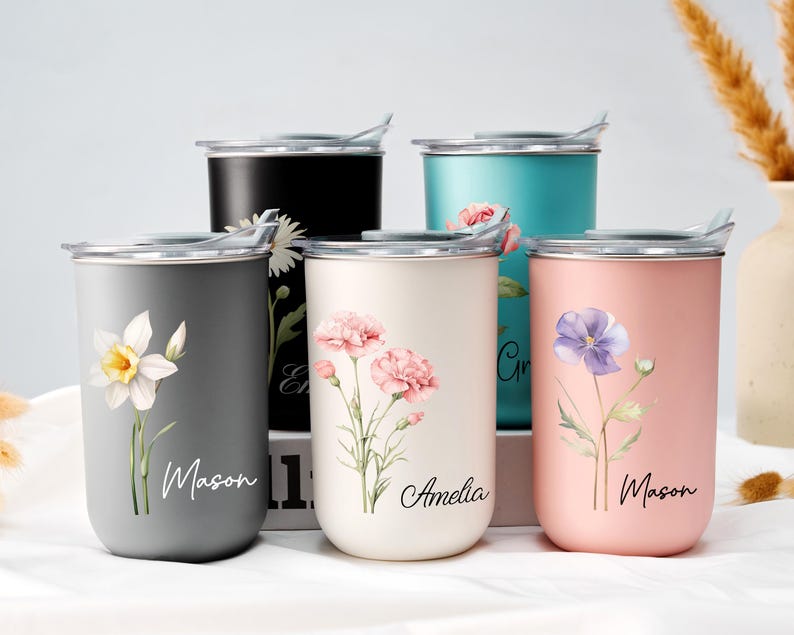 Op de afbeelding: Een verzameling ge&iuml;soleerde bekers in verschillende kleuren, waaronder grijs, zwart, blauwgroen, cr&egrave;me en roze. Elke beker heeft een bloemenontwerp en een naam in script. De bekers hebben zilveren deksels en zijn ontworpen voor dranken.