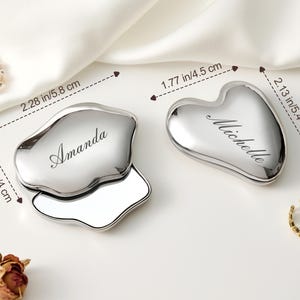 May include: Two silver compact mirrors. One is an irregular shape, 2.28 in (5.8 cm) long, with "Amanda" engraved. The other is heart-shaped, 2.13 in (5.4 cm) long, with "Michelle" engraved. Both have a mirror inside.