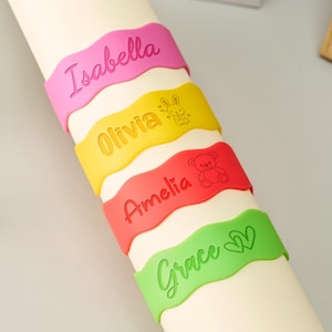 May include: Four wavy silicone wristbands in pink, yellow, red, and light green. The pink band features the name Isabella. The yellow band features the name Olivia and a rabbit icon. The red band features the name Amelia and a teddy bear icon. The green band features the name Grace and two heart icons.