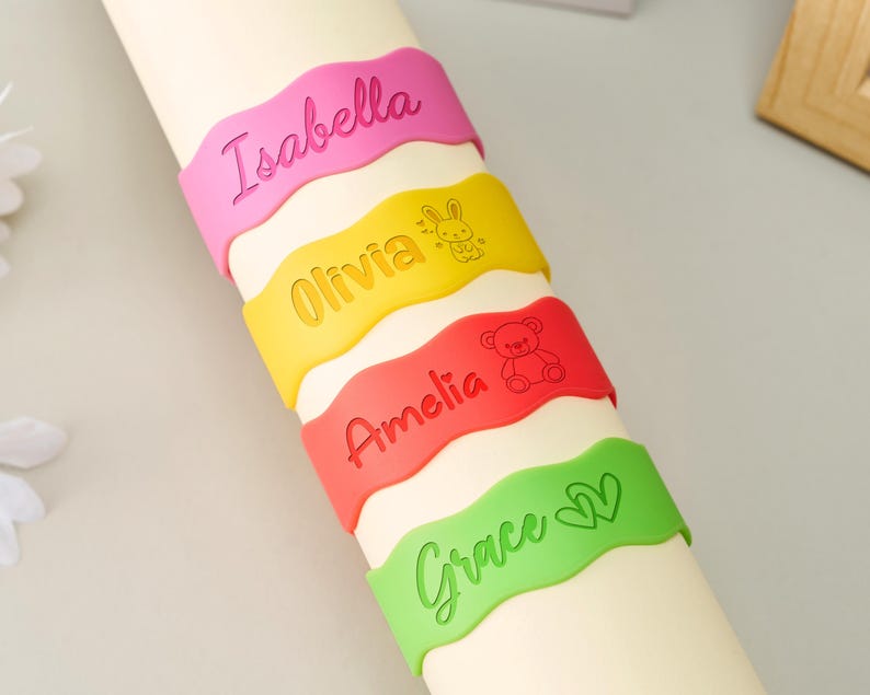 May include: Four wavy silicone wristbands in pink, yellow, red, and light green. The pink band features the name Isabella. The yellow band features the name Olivia and a rabbit icon. The red band features the name Amelia and a teddy bear icon. The green band features the name Grace and two heart icons.