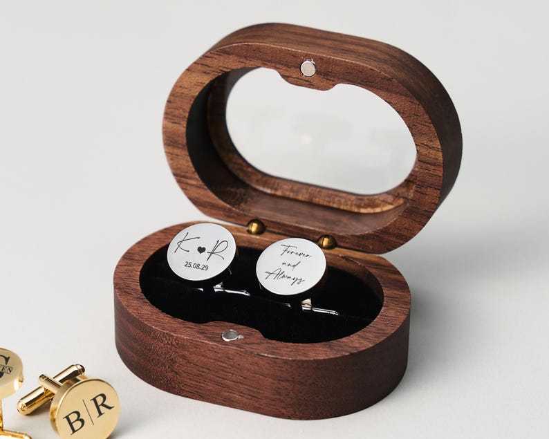 May include: A wooden oval-shaped box with a clear top, holding two silver cufflinks. One cufflink has the initials "K.R" and the date "25.08.29". The other cufflink says "Forever and Always".