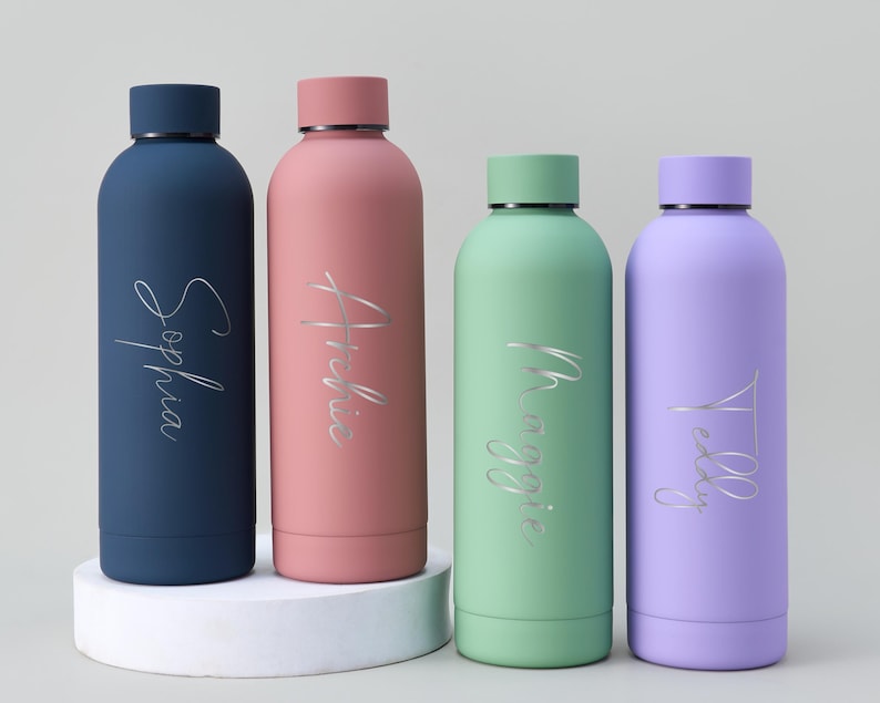 May include: Four colorful, personalized water bottles. The bottles are navy blue, pink, mint green, and lavender. Each bottle has a silver script name: Sophia, Archie, Maggie, and Teddy. The bottles are cylindrical with matching colored lids.