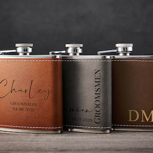 May include: Three personalised hip flasks with silver tops. The flasks are in shades of brown and grey, with stitched leather-effect exteriors. One flask has the name "Charley" and the date "04-08-2025". Another says "Groomsmen" and a date. The third has the initials "DM".