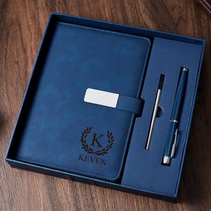 May include: A blue leather journal and pen set in a presentation box. The journal has a silver clasp and is embossed with a letter "K" inside a laurel wreath and the name "Keven". The set includes a pen and a stylus.