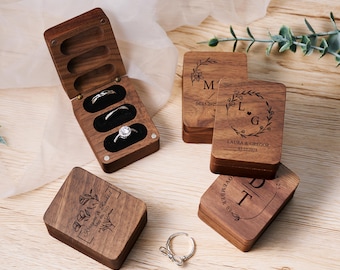 May include: Four wooden ring boxes with engraved floral designs and personalized text. The boxes are made of dark wood and have a natural finish. The text on the boxes includes names, dates, and initials.