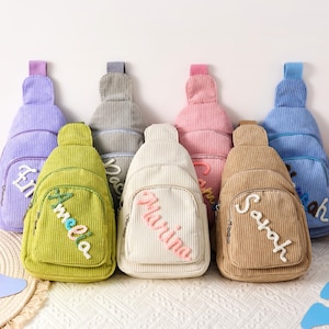 Könnte beinhalten: Eine Auswahl an Cord-Umhängetaschen in verschiedenen Farben: Lila, Grün, Creme, Rosa, Braun und Blau. Jede Tasche ist mit einem gestickten Namen personalisiert. Die Taschen haben eine vertikale Rippenstruktur und einen einzelnen Schultergurt.
