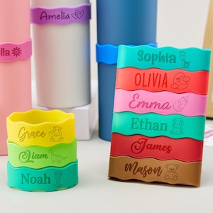 May include: A collection of colorful silicone bands with wavy edges, each personalized with a name and a small icon. The bands are displayed in stacks and wrapped around cylindrical containers, featuring names like Isabella, Amelia, Sophia, Olivia, Emma, Ethan, James, Mason, Grace, Liam, and Noah.