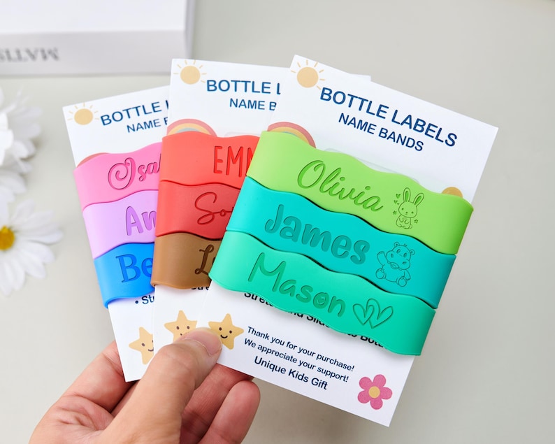 May include: Three sets of colorful silicone bottle labels or name bands featuring wavy edges. The bands are displayed on white cards labeled BOTTLE LABELS NAME BANDS. The bands are printed with various names like Olivia, James, and Mason, along with small icons including a rabbit, a hippo, and hearts.