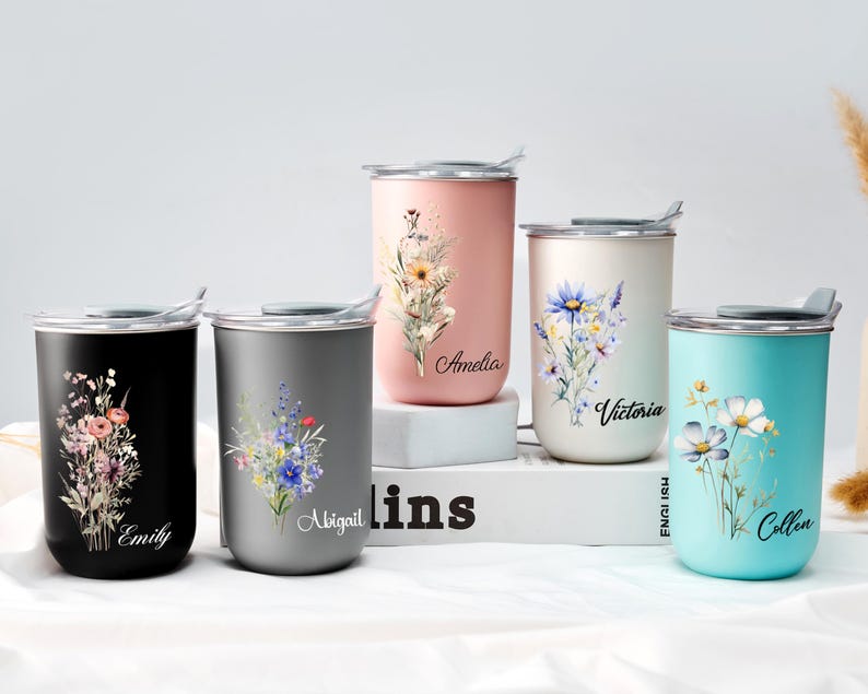 Op de afbeelding: Vijf roestvrijstalen reisbekers in zwart, grijs, roze, cr&egrave;me en turquoise. Elke beker heeft een bloemenontwerp en een naam: Emily, Abigail, Amelia, Victoria en Colleen. De bekers hebben zilveren deksels en staan op een wit oppervlak.