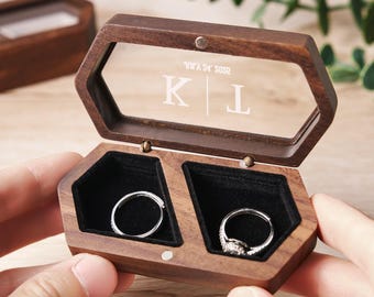 May include: A wooden ring box with a clear lid and two compartments. The lid has the initials "K & L" engraved on it. The box is open and two rings are visible inside.
