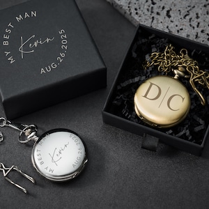 May include: A silver pocket watch with a chain and clasp, alongside a black box inscribed with "MY BEST MAN Kevin AUG.26.2025". A gold pocket watch with the initials "D|C" is presented in a black box.