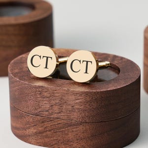 May include: Gold-tone cufflinks with the initials "CT" in black lettering, resting on a dark brown wooden box. The cufflinks are round and shiny, with a classic design. The box has a smooth, polished finish.