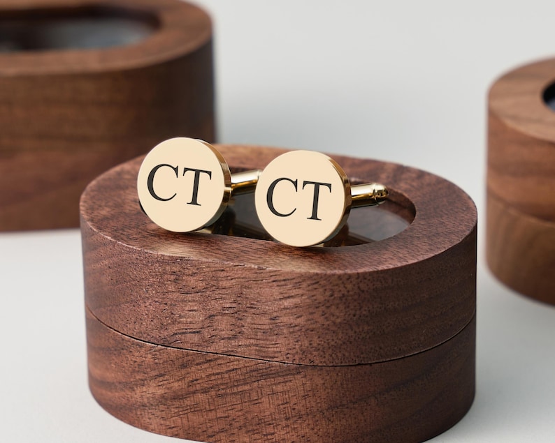 May include: Gold-tone cufflinks with the initials "CT" in black lettering, resting on a dark brown wooden box. The cufflinks are round and shiny, with a classic design. The box has a smooth, polished finish.