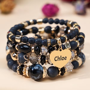 Op de afbeelding: Een stapel armbanden met zwarte, grijze en gouden kralen. De armbanden hebben verschillende kralenmaten en -vormen, met een gouden hartvormige bedel met de naam "Chloe".