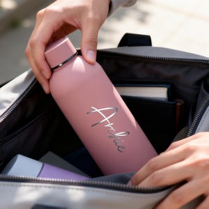 May include: A pink water bottle with the name "Archie" in silver script is being placed into a gray bag. The bottle has a matching pink cap with a black band. The bag contains other items, including a book and a purple bottle.