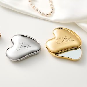 May include: Two heart-shaped compact mirrors, one silver and one gold, are displayed on a white surface. The silver compact has the name "Jennifer" inscribed, while the gold compact has "Stephanie." A pearl necklace and a dried rose are also present.