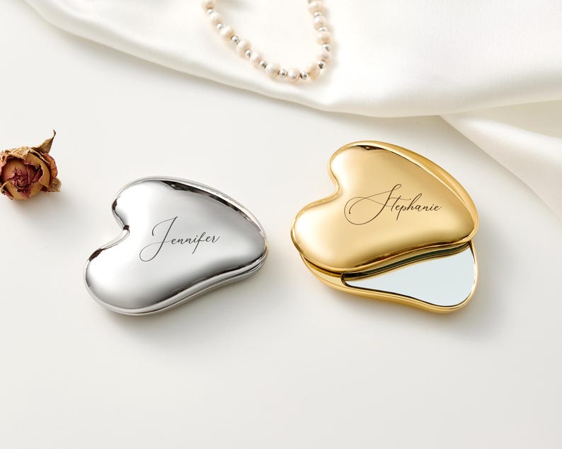 May include: Two heart-shaped compact mirrors, one silver and one gold, are displayed on a white surface. The silver compact has the name "Jennifer" inscribed, while the gold compact has "Stephanie." A pearl necklace and a dried rose are also present.