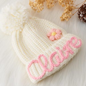 Può includere: Berretto di lana bianco con un pon pon rosa e un fiore rosa. Il berretto ha il nome "Claire" ricamato in filo rosa.