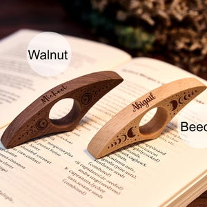 May include: Two wooden thumb page holders, one walnut and one beech, rest on an open book. The walnut holder is engraved with "Michael" and celestial designs. The beech holder is engraved with "Abigail" and similar celestial designs.