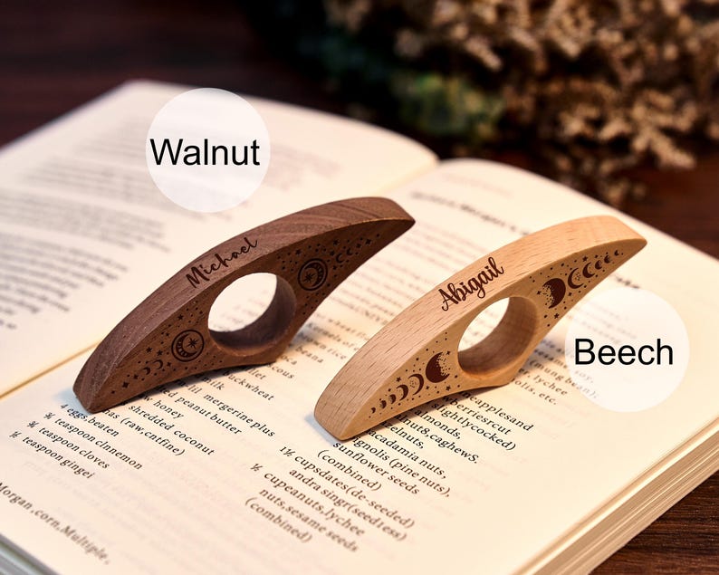 May include: Two wooden thumb page holders, one walnut and one beech, rest on an open book. The walnut holder is engraved with "Michael" and celestial designs. The beech holder is engraved with "Abigail" and similar celestial designs.
