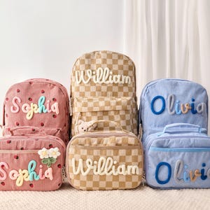 May include: Three sets of corduroy backpacks and lunch bags in pink with strawberry prints, tan checkered patterns, and light blue stripes. Each item is personalized with raised, colorful fabric lettering reading Sophia, William, or Olivia.