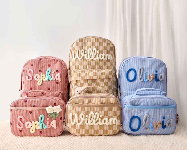 May include: Three sets of corduroy backpacks and lunch bags in pink with strawberry prints, tan checkered patterns, and light blue stripes. Each item is personalized with raised, colorful fabric lettering reading Sophia, William, or Olivia.
