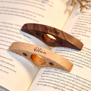 May include: Two wooden thumb page holders with engraved names and celestial designs. One is light-colored, the other dark. The names "William" and "Olivia" are etched on each. They are resting on an open book.