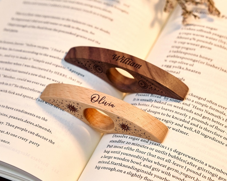 May include: Two wooden thumb page holders with engraved names and celestial designs. One is light-colored, the other dark. The names "William" and "Olivia" are etched on each. They are resting on an open book.