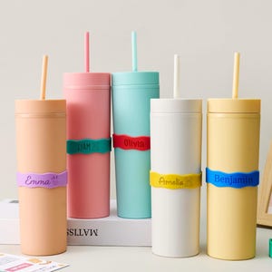 May include: A collection of personalized tumblers in various pastel colors, each with a matching straw. Each tumbler has a colorful band with a name on it: Emma, Liam, Olivia, Amelia, and Benjamin. The tumblers are cylindrical and sit on a white surface.