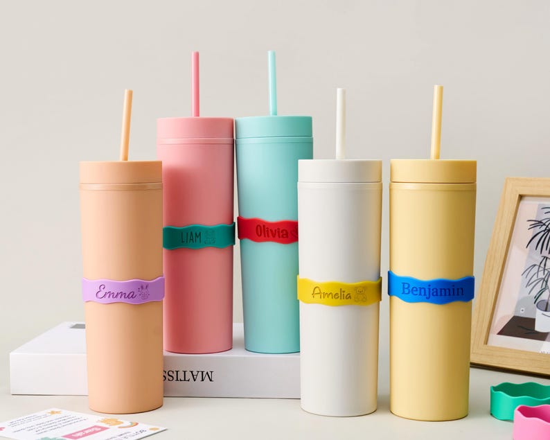 May include: Five cylindrical tumblers in peach, pink, teal, white, and yellow, each with a matching straw and a wavy silicone band. The bands are personalized with the names Emma, Liam, Olivia, Amelia, and Benjamin, along with small icons.