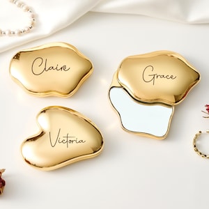 May include: Gold-colored compact mirrors with personalized names: Claire, Grace, and Victoria. One mirror is open, revealing a reflective surface. The mirrors are on a white surface with a pearl necklace, a ring, and dried flowers.