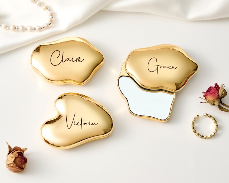 May include: Gold-colored compact mirrors with personalized names: Claire, Grace, and Victoria. One mirror is open, revealing a reflective surface. The mirrors are on a white surface with a pearl necklace, a ring, and dried flowers.