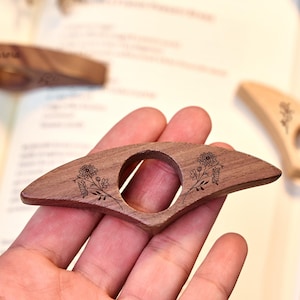 May include: A wooden book page holder with a floral design. The dark brown, fan-shaped holder has a circular opening and engraved flowers. Other similar holders are visible in the background, resting on an open book.
