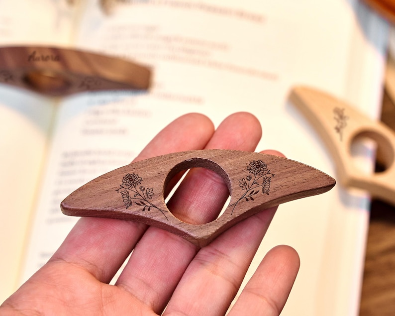 May include: A wooden book page holder with a floral design. The dark brown, fan-shaped holder has a circular opening and engraved flowers. Other similar holders are visible in the background, resting on an open book.