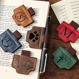 Può includere: Portapenne in pelle di vari colori, ognuno con un design unico e fascia elastica. I disegni includono un'iniziale, un'impronta di zampa, un nome, una croce con una colomba e una ghirlanda con un'iniziale. Una penna è al centro.