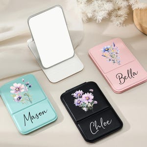 May include: A collection of personalized compact mirrors in various colors. One is open, showing a white mirror. Others are pink, teal, and black, each adorned with floral designs and names: Bella, Mason, and Chloe. The mirrors are on a neutral surface.