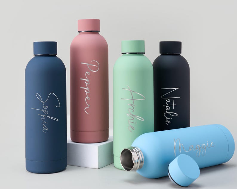 May include: A collection of personalized water bottles in various colors, including blue, pink, green, and black. Each bottle features a name in a silver script font. One bottle is lying on its side with the cap removed.
