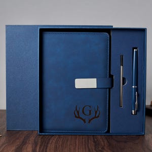 Op de afbeelding: Een blauwe leren notitieboek- en pennenset in een geschenkverpakking. Het notitieboek heeft een zilveren sluiting en een reliëf hertengeweimotief met initialen. De pen en de vulling zijn ook blauw. De doos is open.