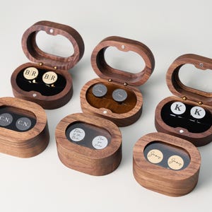May include: Wooden cufflink boxes with clear oval lids. Each box contains a pair of cufflinks with initials or names. The boxes are open, revealing the cufflinks inside. The cufflinks are gold and silver.