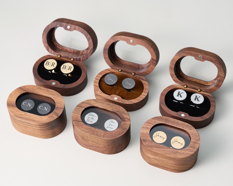 May include: Wooden cufflink boxes with clear oval lids. Each box contains a pair of cufflinks with initials or names. The boxes are open, revealing the cufflinks inside. The cufflinks are gold and silver.
