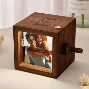 May include: A dark brown wooden photo cube with a rotating mechanism, displaying a couple's photo. The cube has the words "Love You More" engraved on top. A small wooden handle is on the side.