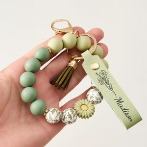 May include: A beaded keychain bracelet in shades of green, cream, and gold. The bracelet features large round beads, decorative floral beads, and a tassel. A light green leather tag with the name "Madison" and a floral design is attached.